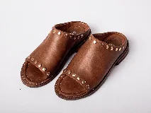 Sandalias de cuero color marrón claro, con punta descubierta y diseño tipo mule. Presentan tachas doradas en el borde superior y suela plana con pequeño tacón. Fabricadas en Uruguay.
