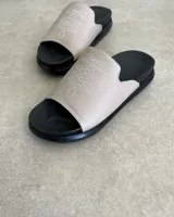 Sandalias chatas color beige con base negra y detalle de bordado blanco en la tira superior.