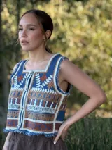 Chaleco corto de denim azul con bordado artesanal en tonos naranja, blanco y marrón. Tiene escote cuadrado, cierre de botones y terminaciones desflecadas.