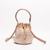 Bolso tipo saco o bucket bag, con cuerpo tejido en rafia color beige y detalles en cuero sintético color camel. Posee un asa superior corta y una correa larga ajustable para llevar al hombro o cruzado. El cierre es ajustable mediante cordón.