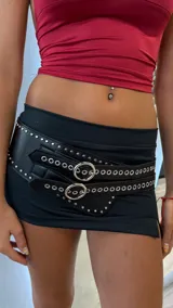 Cinturón ancho de cuero sintético negro con doble tira, múltiples ojales y tachas metálicas redondas. Presenta dos hebillas metálicas circulares grandes en el frente.