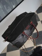 Bolso de viaje tipo weekender de cuero color marrón con detalles en rojo en los cierres. Tiene doble asa corta y correa de hombro ajustable y extraíble. Cuenta con bolsillos internos y externos.