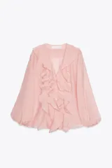 Camisa color rosa pálido confeccionada en tejido ligero con lyocell. Presenta escote en V, cierre frontal con botones, mangas largas abullonadas con puños elásticos y detalle de volantes en el frente.