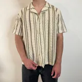 Camisa de manga corta con cuello solapa y cierre abotonado. Tejido calado con rayas verticales en tonos beige y marrón.