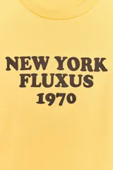 Camiseta amarilla de algodón con cuello redondo, manga corta y estampado marrón con el texto "New York Fluxus 1970".