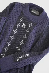 Sweater de punto azul marino con cuello redondo y mangas largas. Presenta textura trenzada en los laterales y detalle de estrellas bordadas en blanco en el centro, con la inscripción "Del Sur" en la parte superior.