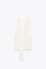 Chaleco largo color blanco, de escote en pico y sin mangas. Tiene bolsillos de vivo en el delantero, aberturas laterales y cinturón multiposición del mismo tejido.