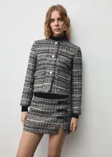 Conjunto de chaqueta y falda de tweed en tonos blanco, negro y dorado. La chaqueta es corta con cuello redondo, botones metálicos y bolsillos de parche. La falda es mini con abertura lateral y detalle de lazos.