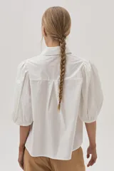 Camisa blanca de mangas abullonadas con puños elásticos, cuello clásico y cierre frontal con botones. Presenta detalles de bordado calado en el pecho.