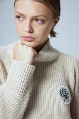 Sweater de punto grueso color crema, con cuello alto y parche circular con bordado en el pecho.