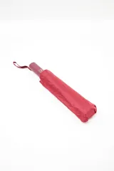 Paraguas plegable de color rojo oscuro o bordó, con funda de tela a juego y correa de transporte. El mango es del mismo color y tiene un pequeño logo blanco.