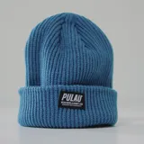 Gorro de lana tejido, color celeste, con etiqueta negra con el logo de la marca Pulau en el frente.