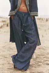Pantalón ancho azul con textura de lino, tiro alto, cintura elástica con cordón ajustable, pinzas delanteras, bolsillos laterales y traseros, y cinturón de la misma tela con apliques metálicos.