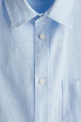 Camisa celeste de lino y algodón, corte regular, manga larga, cuello inglés, bolsillo en el pecho y cierre con botones.