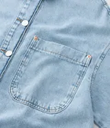 Camisa de jean de algodón, color celeste claro lavado, con cuello tradicional, manga larga y bolsillo de parche en el pecho. Presenta un cierre frontal con botones metálicos.