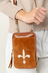 Bolso tipo pocket bag de cuero vacuno color marrón, con bordado de flor de lis en color claro en el frente. Posee cierre principal, bolsillo trasero y correa desmontable.