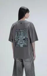 Remera de algodón premium color gris oscuro, corte oversize con hombros caídos y cuello redondo. Estampado grande en la espalda con la palabra "KILOMBITO" sobre una ilustración de una figura en un entorno boscoso en tonos verde azulado y negro.