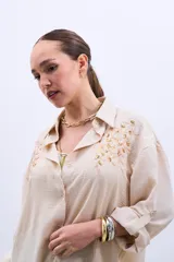 Camisa color beige con bordado de lentejuelas doradas en los hombros, corte clásico, cuello camisero y mangas largas con puños abotonados.