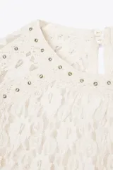 Top de encaje color blanco, con cuello redondo, mangas largas acampanadas y detalle de tachas metálicas en cuello y hombros. Cierre en la espalda con abertura y botón.