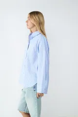 Camisa celeste con finas rayas verticales negras, de corte amplio, manga larga, cuello clásico, cierre frontal con botones y puños abotonados.