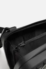 Bandolera rígida negra con acabado engomado y relieve. Tiene un compartimento principal con doble cremallera, un bolsillo interior de rejilla y asa de hombro ajustable y extraíble.