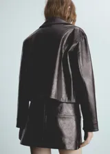 Chaqueta corta de poliuretano color marrón oscuro con textura, cuello camisero, manga larga con puños abotonados, dos bolsillos de solapa en la parte delantera y cierre frontal con botones.