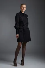 Vestido camisero corto negro con cuello, cierre frontal con botones dorados, cinturón con hebilla circular y mangas largas abullonadas con puños abotonados. Presenta bolsillos con solapa en el pecho y en la falda.