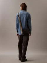 Bolso bandolera rectangular de poliuretano texturizado color negro, con cierre de cremallera superior y logo de Calvin Klein grabado en relieve en la parte frontal inferior. Posee una correa ancha de tela ajustable en tono verde oliva/grisáceo.