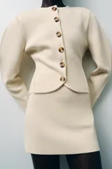 Chaqueta color crema de punto con cuello redondo, manga larga, corte entallado, bajo asimétrico y cierre frontal con botones marrones.