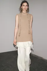 Vestido color beige, de corte recto, con cuello redondo y sin mangas. Presenta un tejido de punto en la parte superior y un volante de encaje blanco en el ruedo.