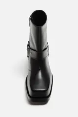 Bota negra de cuero con caña corta, horma cuadrada, suela a tono con ligero tacón y arnés decorativo a la altura del tobillo con detalles metálicos.