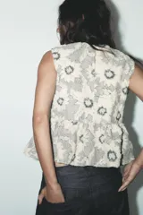 Top sin mangas color crudo con estampado de flores negras, cuello redondo, peplum en la cintura y cierre trasero con cremallera metálica.