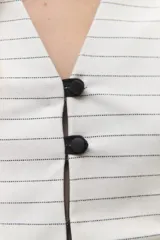 Chaleco blanco con finas rayas verticales negras, escote en V, cierre frontal con botones negros y volados en lino negro en los hombros. La parte inferior presenta otro volado en la misma tela del cuerpo.