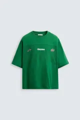 Camiseta verde con corte holgado, cuello redondo y mangas cortas. Presenta una banda horizontal en el pecho y la espalda con tejido de red. Estampados combinados a contraste en el frente.