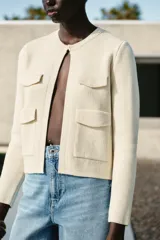 Chaqueta color crema de cuello redondo y manga larga, con bolsillos de parche con solapa en el frente y cierre frontal con corchete oculto.