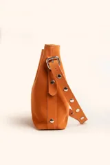 Cartera tipo balde de gamuza color marrón, con correa ajustable decorada con tachas metálicas plateadas y logo grabado en la parte frontal.