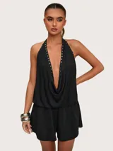 Mono corto negro con escote halter profundo y drapeado, adornado con tachas metálicas plateadas a lo largo del escote. La cintura es elástica y la parte inferior es de estilo short.