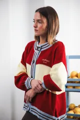 Cardigan tejido a rayas horizontales rojas y crema, con cuello redondo, cierre frontal con botones marrones y mangas largas abullonadas. Presenta ribetes a rayas en cuello, puños y ruedo. Tiene bordado el logo de la marca en el pecho.