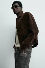 Cazadora de hombre de corte relajado confeccionada en piel con acabado ante, color marrón oscuro. Presenta cuello solapa, manga larga con puños abotonados a presión, bolsillos de vivo en cadera, bolsillo interior y bajo con elástico. Cierre frontal oculto por solapa con botones a presión.