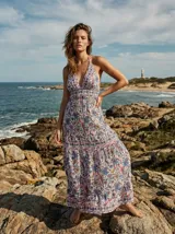 Vestido largo con estampado floral y de aves en tonos rojizos y azules sobre fondo crema. Presenta escote halter profundo en V y cintura marcada con falda con vuelo en capas.