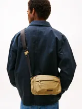 Bandolera Tommy Hilfiger color beige con logo texturizado en el frente. Tiene correa ajustable de tejido trenzado, cierre de cremallera y bolsillo exterior con cremallera.