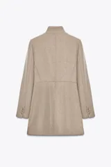 Abrigo color beige confeccionado en lana. Tiene cuello subido, mangas largas con hombreras, forro interior combinado y cierre frontal cruzado con botones metálicos.