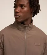 Campera deportiva color marrón, con cuello alto, cierre frontal con cremallera, manga larga, bolsillos laterales y detalles de etiqueta aplicada y rayas.