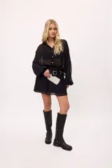 Vestido corto negro de chiffon con transparencia, corte holgado, cuello camisero, mangas largas con puños elásticos, cintura elastizada y cinturón con hebilla cuadrada.