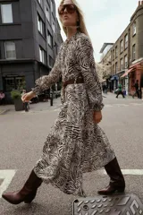 Vestido midi con estampado animal cebra en tonos blanco y negro, cuello redondo con lazada ajustable, manga larga con puños elásticos, forro interior y cinturón marrón con hebilla metálica.