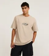 Remera de algodón color beige, corte relaxed (holgado), manga corta y cuello redondo. Presenta un pequeño estampado localizado con lettering estilizado en el pecho.