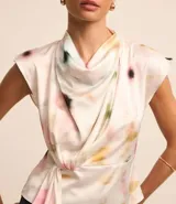 Blusa sin mangas de tela satinada color blanco, con cuello drapeado, lazo lateral y estampado de manchas en tonos rosados, amarillos y verdes.