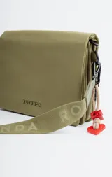 Bandolera color verde oliva con correa de tela ajustable con el nombre de la marca estampado. Incluye un dije colgante con forma de corazón.
