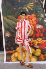 Vestido camisero corto con estampado abstracto en tonos naranja, rosa, amarillo y negro. Tiene cuello camisero, mangas cortas y cierre frontal con botones. Incluye lazo en la cintura para ajustar.