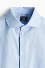 Camisa celeste de algodón con cuello italiano, tapeta clásica y canesú en la espalda. Mangas largas con tapeta y botón, y cierre ajustable de botón en los puños. Bajo levemente redondeado. Corte ajustado que realza el contorno del cuerpo creando una silueta entallada.